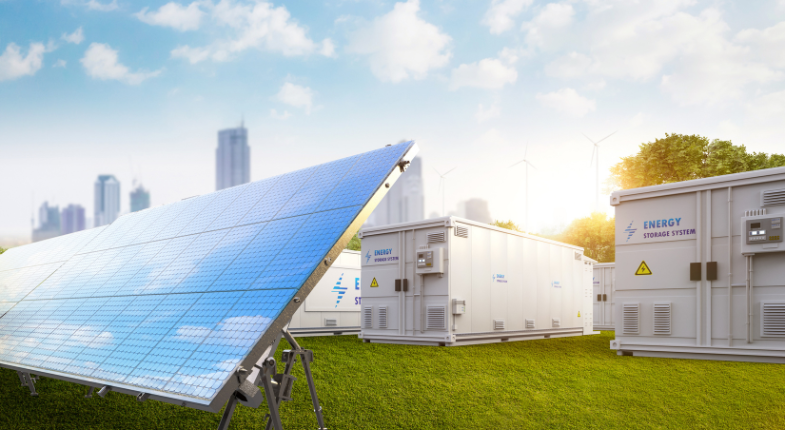 Panel solar en primer plano con sistemas de almacenamiento de energía al fondo, en un entorno verde y con edificios urbanos en el horizonte.